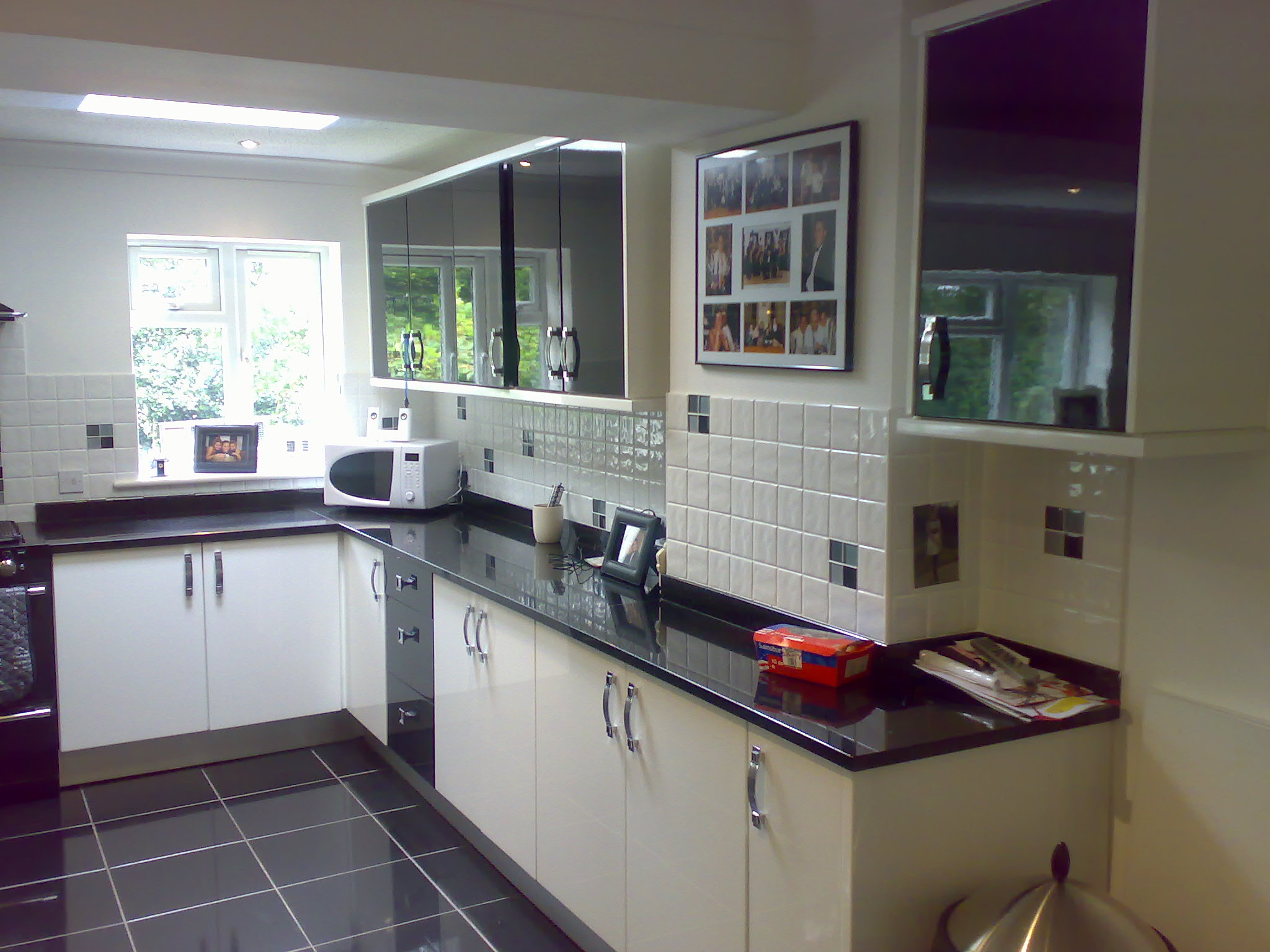 Nowoczesna kuchnia z białymi szafkami dolnymi i czarnymi, połyskującymi szafkami górnymi, czarnym blatem i podłogą w szachownicę, oświetlona światłem dziennym wpadającym przez okno.